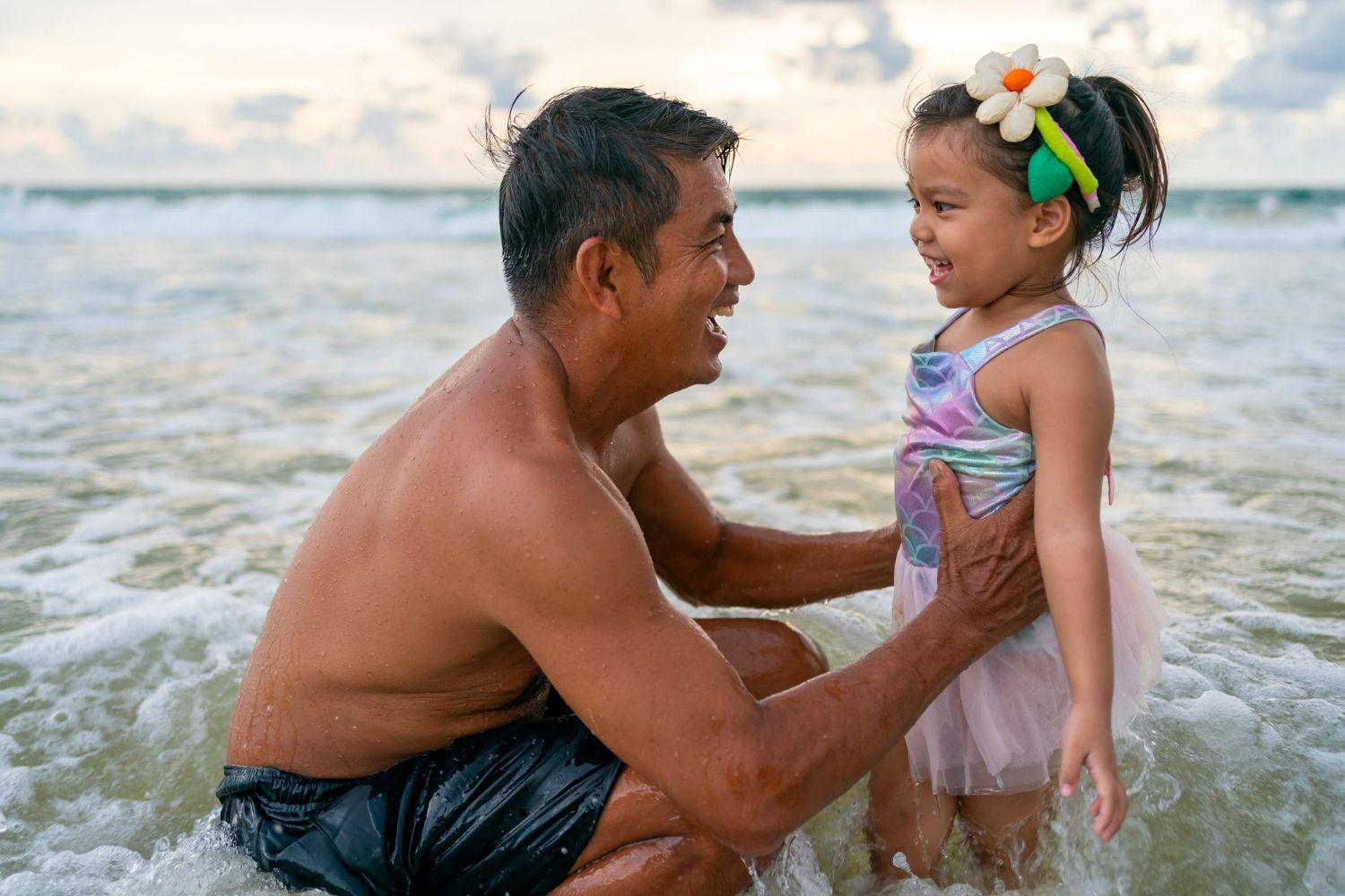 A parent and child smile at each other while playing in the shallow ocean waves at the beach.