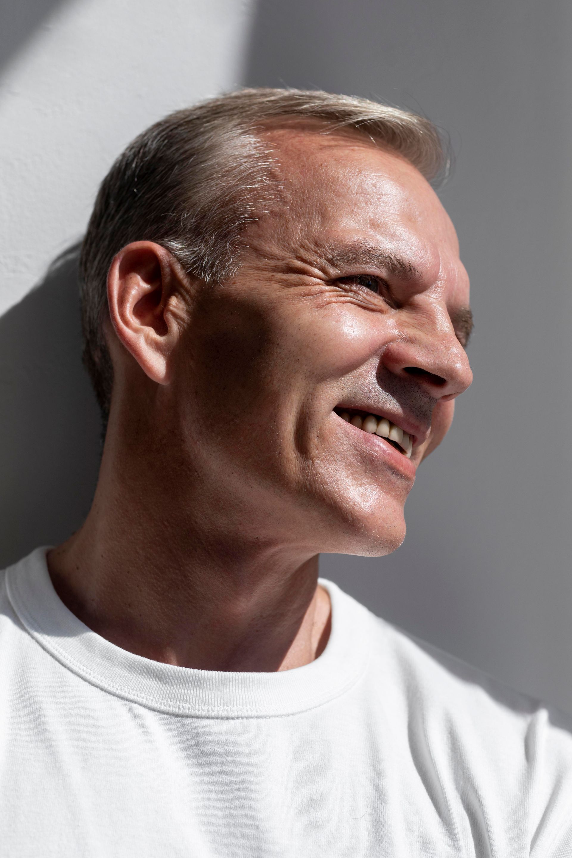 A man in a white shirt is smiling and leaning against a wall.