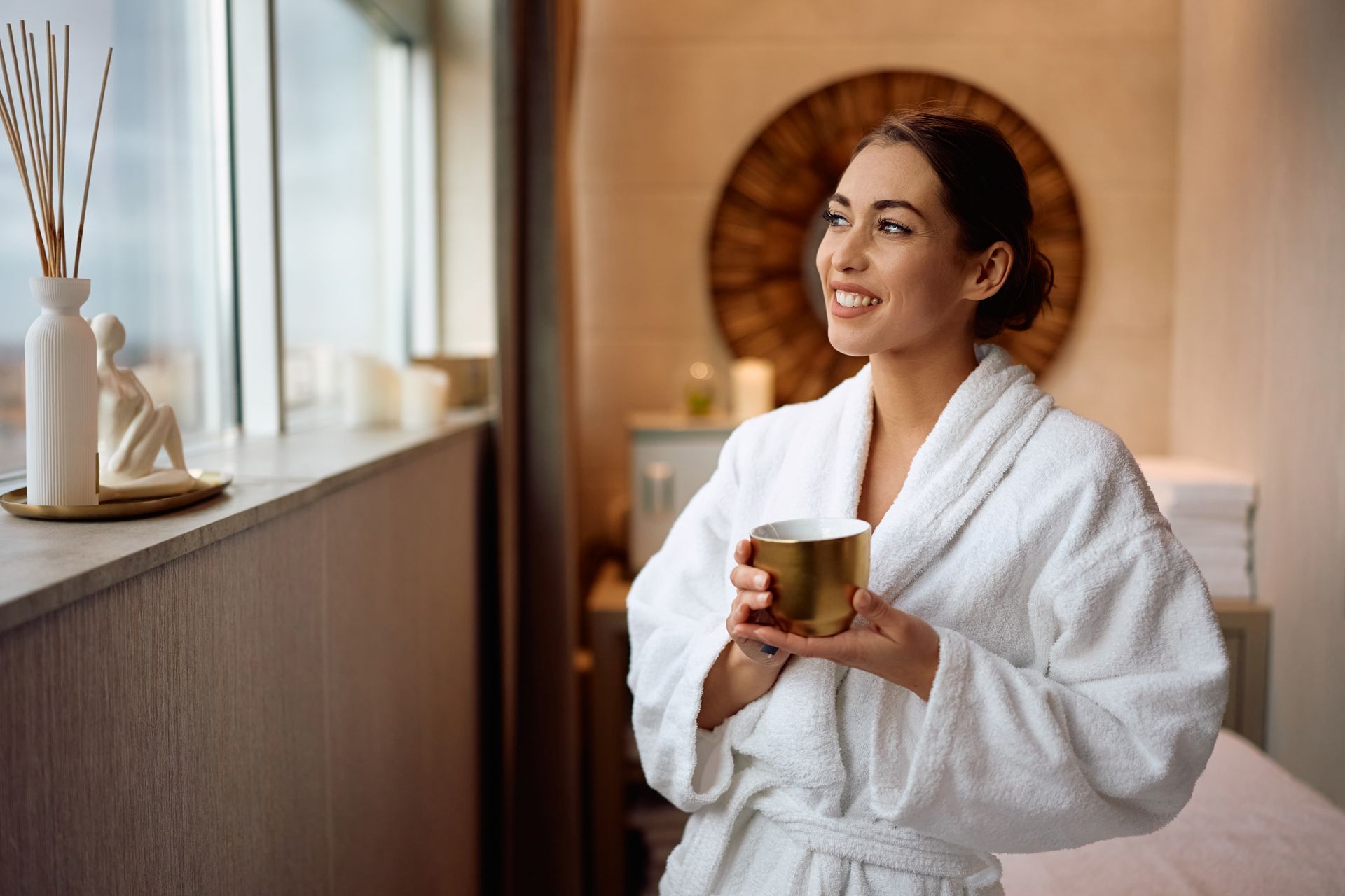 Woman in white robe, smiling, holding a gold cup, looking out a window in a spa-like room.