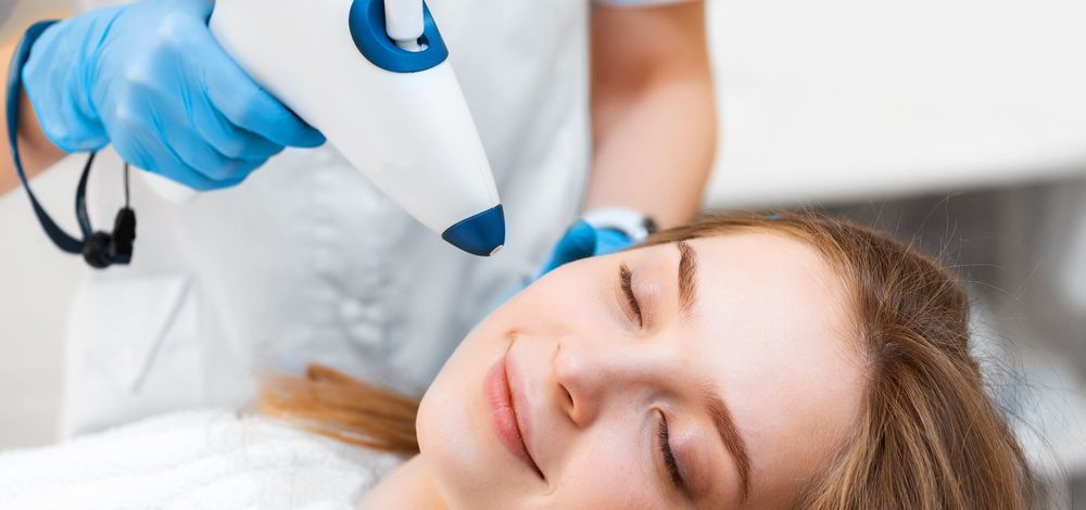 Woman receiving facial laser treatment at a clinic. Her eyes are closed, smiling. Doctor with blue gloves.