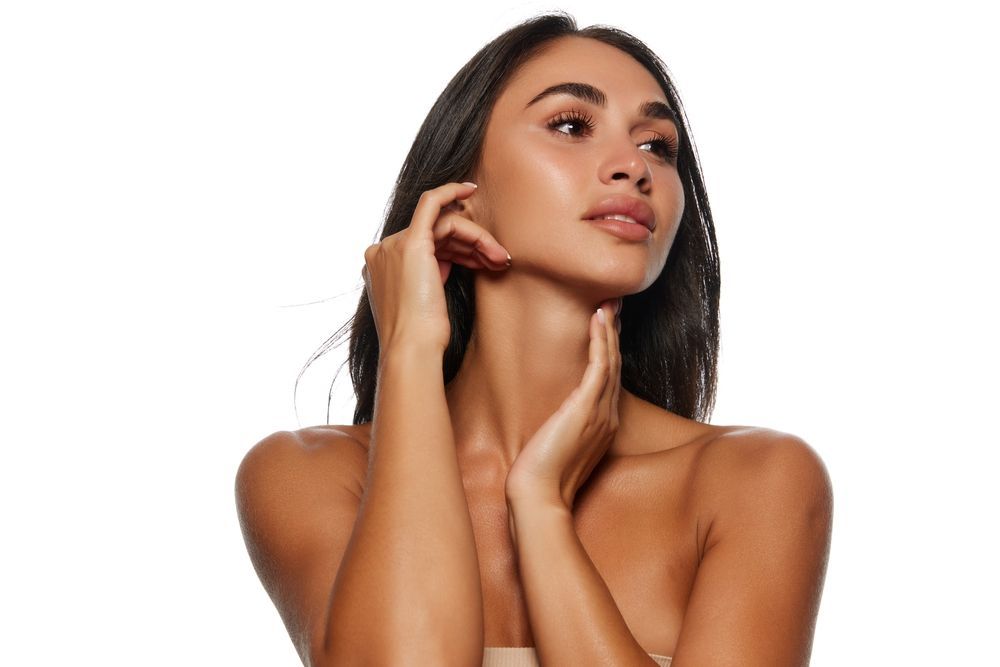 Woman with dark hair, tanned skin, and a serene expression, touching her neck and face with both hands.