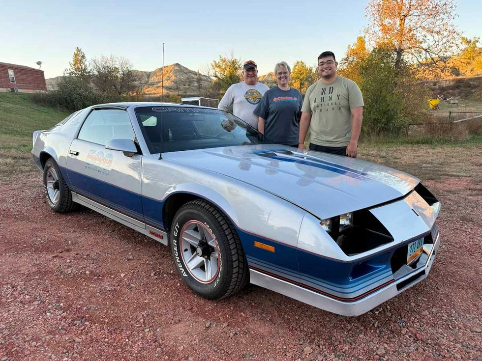 Three men are standing in front of a silver and blue car.