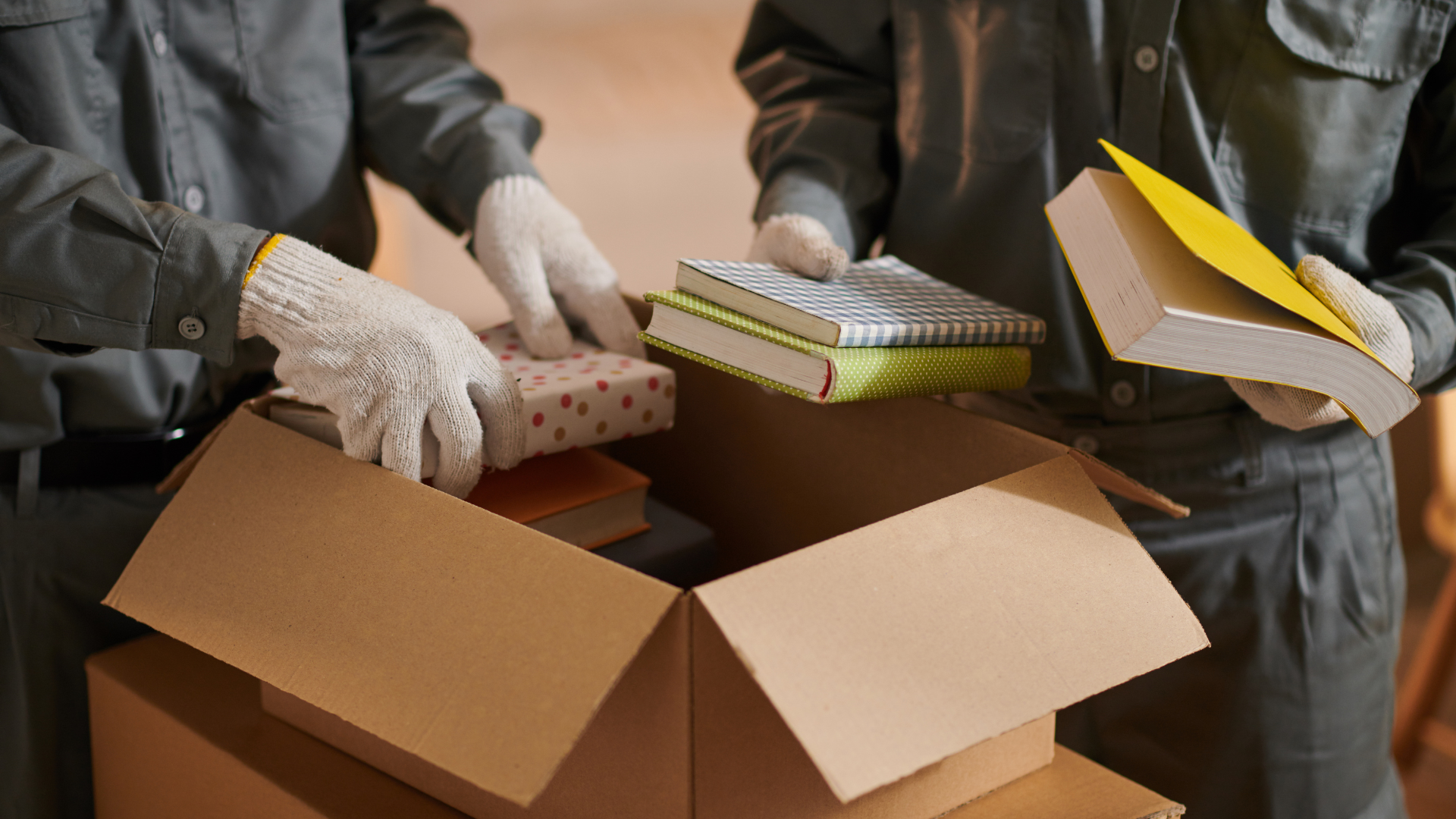 Two people in work clothes, gloves, packing books into cardboard boxes.