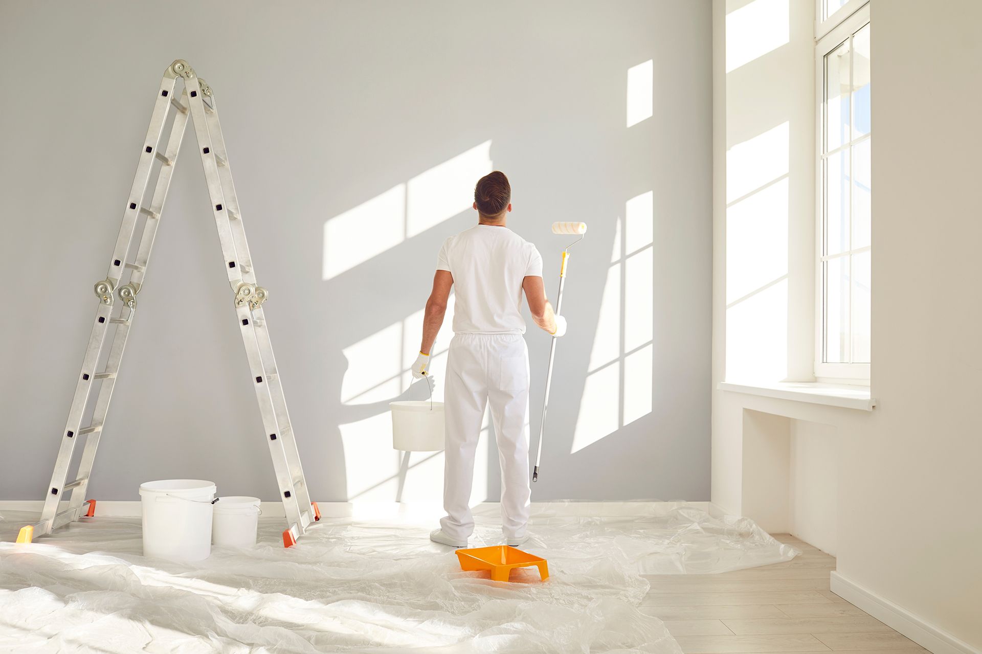 Person painting a gray wall with a paint roller in a room with a ladder and window.