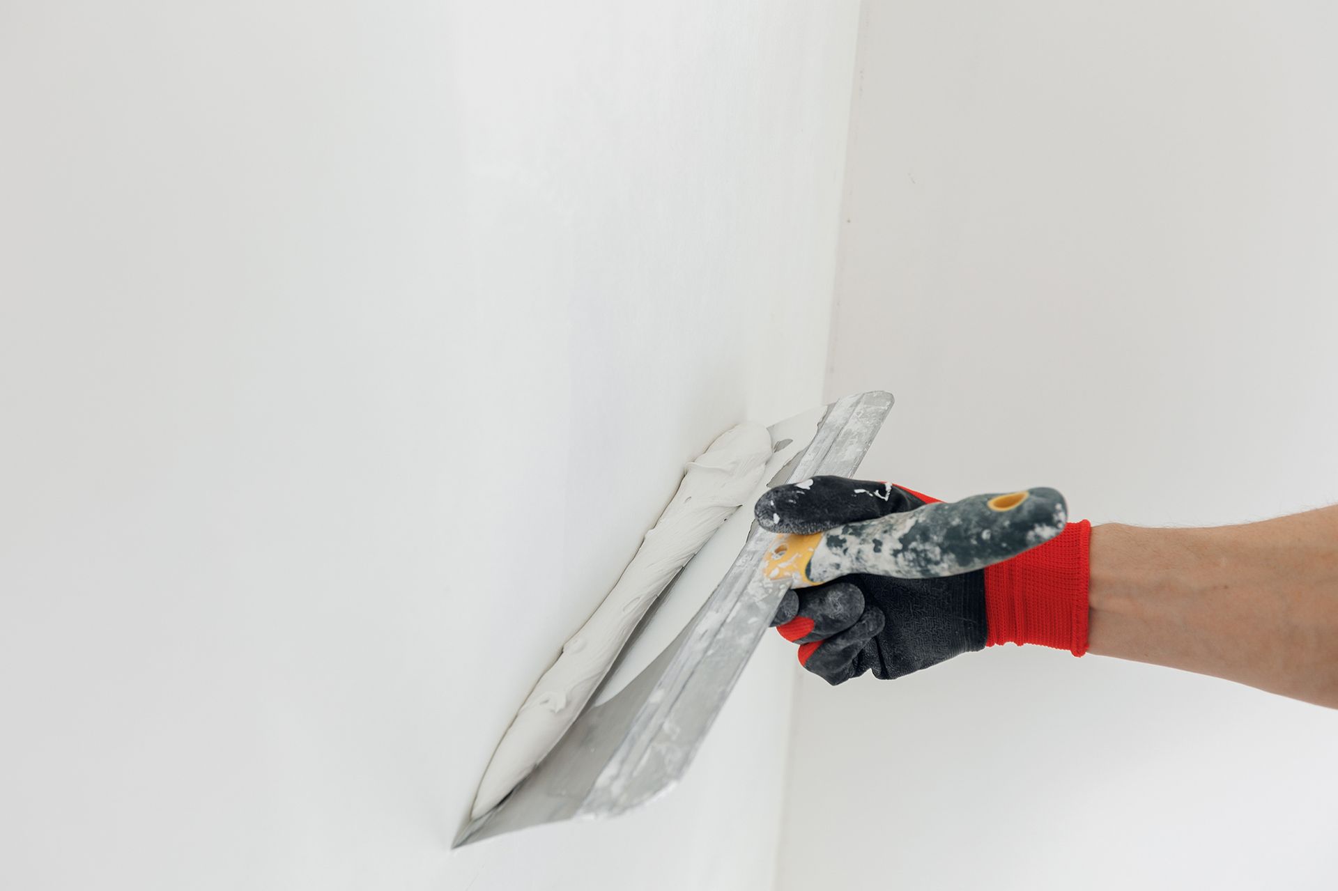 Hand using a trowel to apply drywall mud to a white wall.