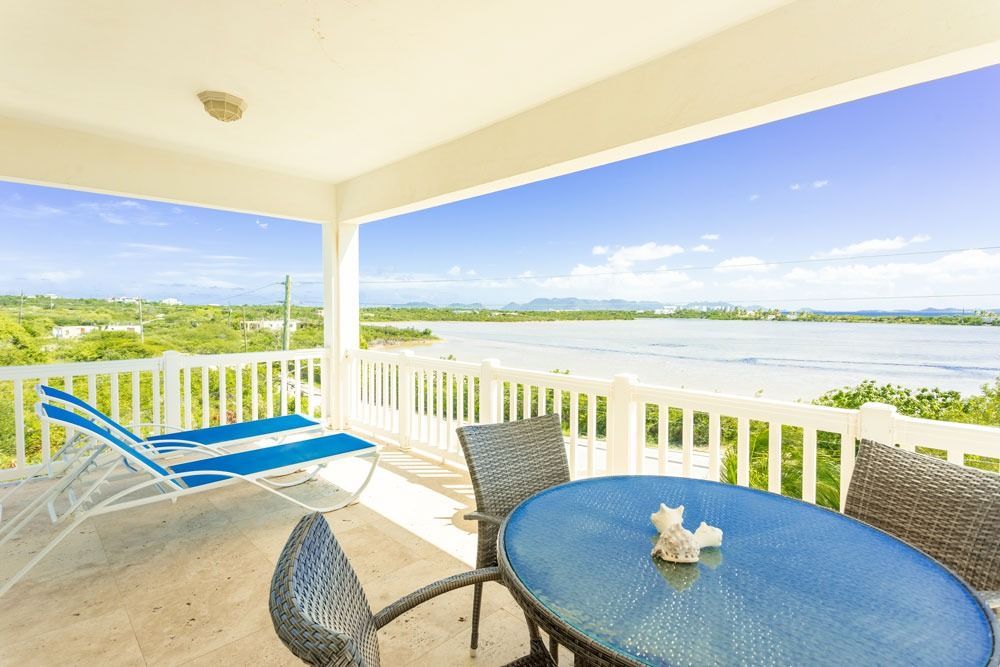 Covered patio overlooking water, featuring a blue table, chairs, and a striped lounge chair.