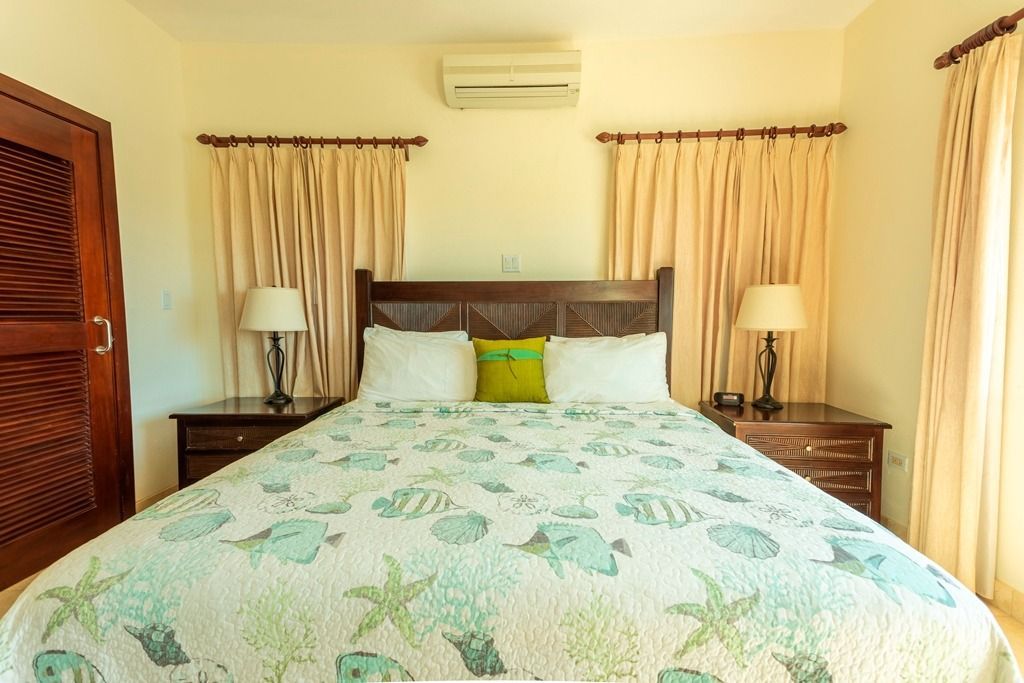Bedroom with a bed, nightstands, lamps, and curtains. Light-colored walls and a wall-mounted AC unit.