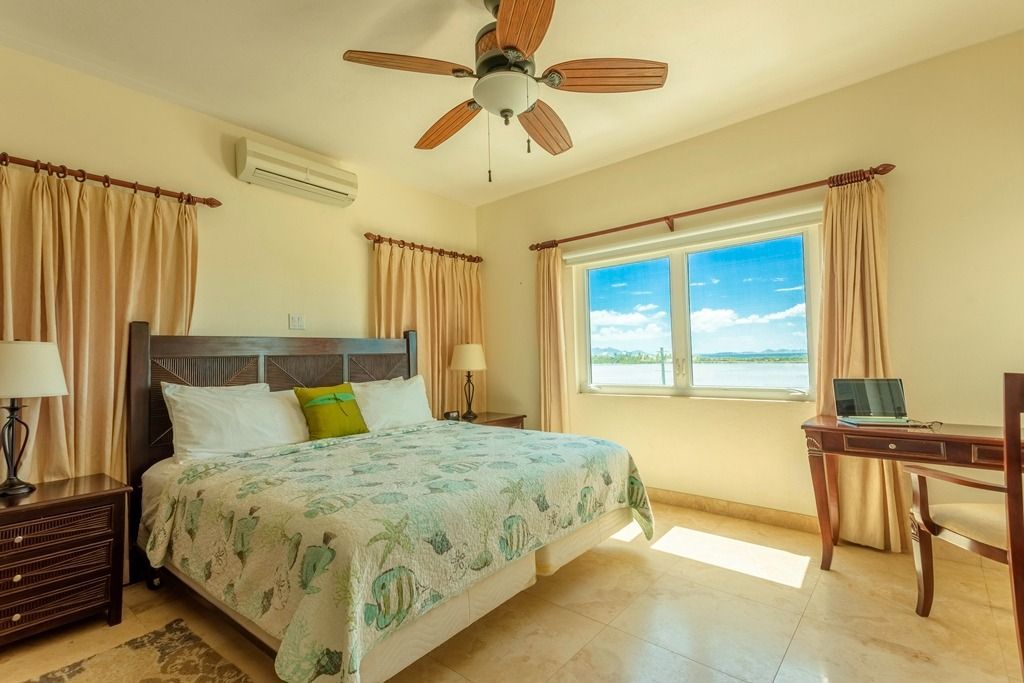 Bedroom with bed, desk, window with sea view, tan curtains and cream-colored walls.