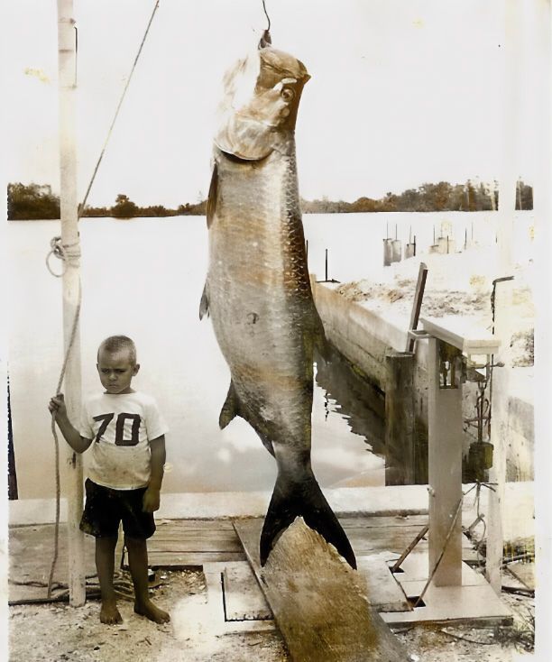 A boy in a 70 shirt stands next to a large fish