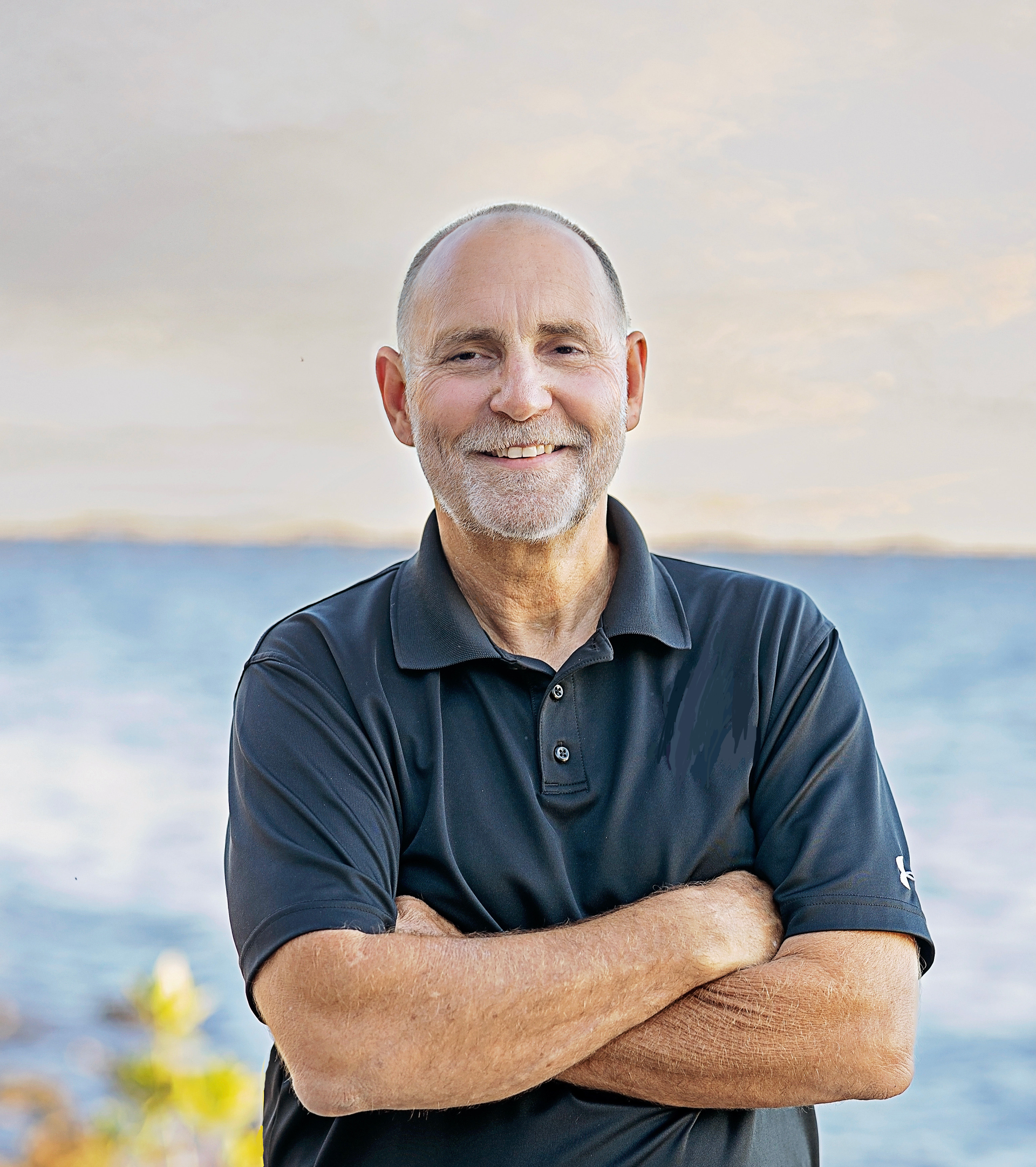 A man with a beard is standing with his arms crossed in front of the ocean.