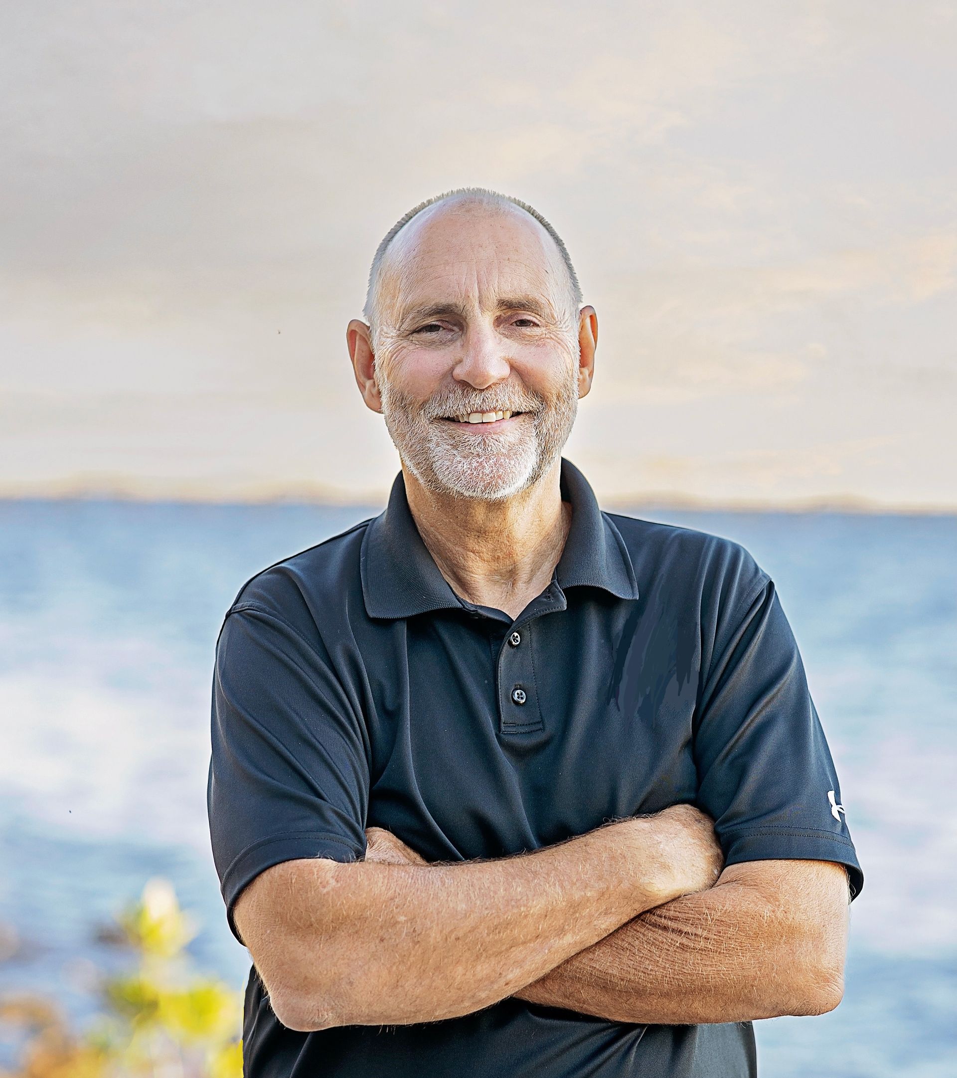 A man with a beard is standing with his arms crossed in front of the ocean.