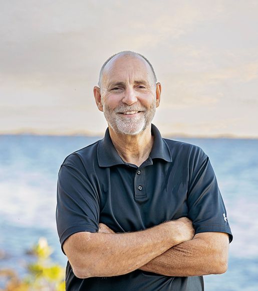 A man with a beard is standing with his arms crossed in front of the ocean.