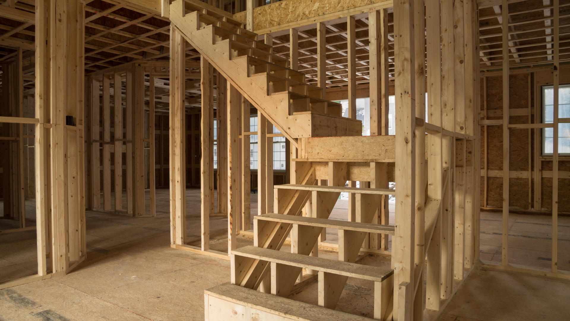 Unfinished interior framing of a house with a staircase. Light wood construction; sawdust on the floor.