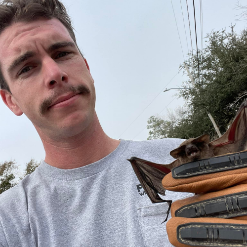Man in gray shirt holding a bat in gloved hands outdoors; overcast sky.
