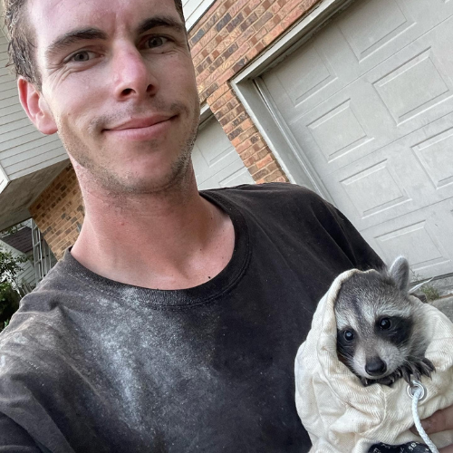 Man holding a raccoon wrapped in a blanket in front of a garage. The raccoon looks at the camera.