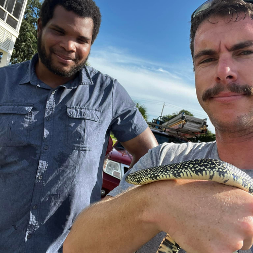 Two men with a snake on one's arm; outdoors under a blue sky. One man smiles; the other holds the snake.