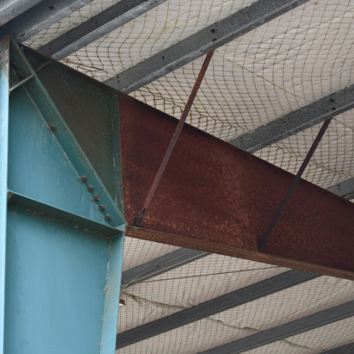Steel beams, rust-colored and blue, supporting a roof structure with a safety net.