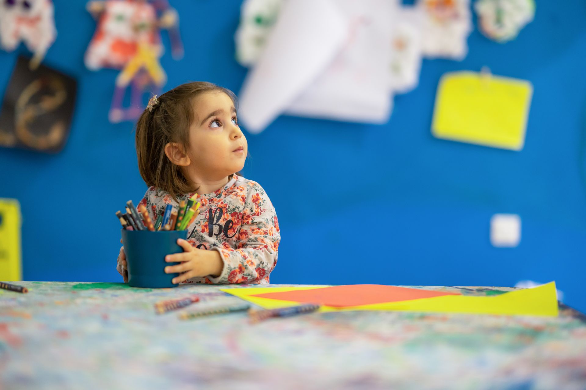 Little girl sitting at a table holding a bucket of pencils and crayons.