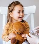 A young girl sitting on a chair holding a teddy bear. The bear is getting a shot and she is smiling.