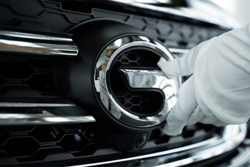 Mão enluvada branca toca o acabamento cromado de uma grade preta de carro com um emblema