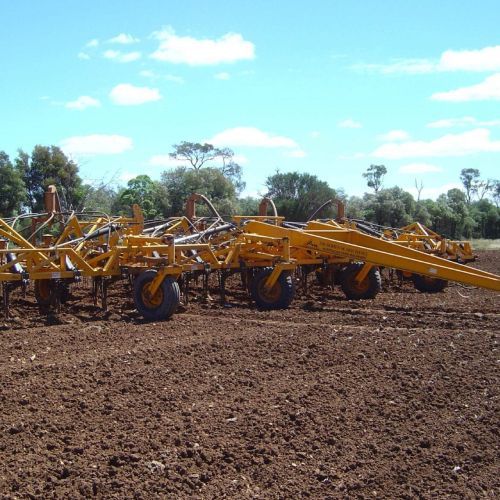 Yellow agricultural cultivator in a field, with trees and blue sky.