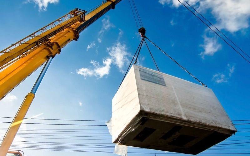 Yellow crane lifting a large, rectangular, wrapped object against a blue sky with power lines.