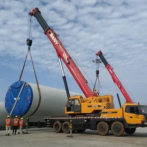 Two red Sany cranes lift a large, blue turbine component at a construction site. Workers in safety vests watch.