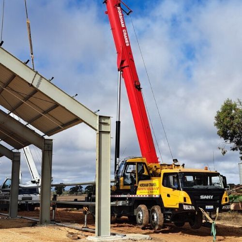 Yellow crane lifting a steel frame at a construction site, under a cloudy sky.
