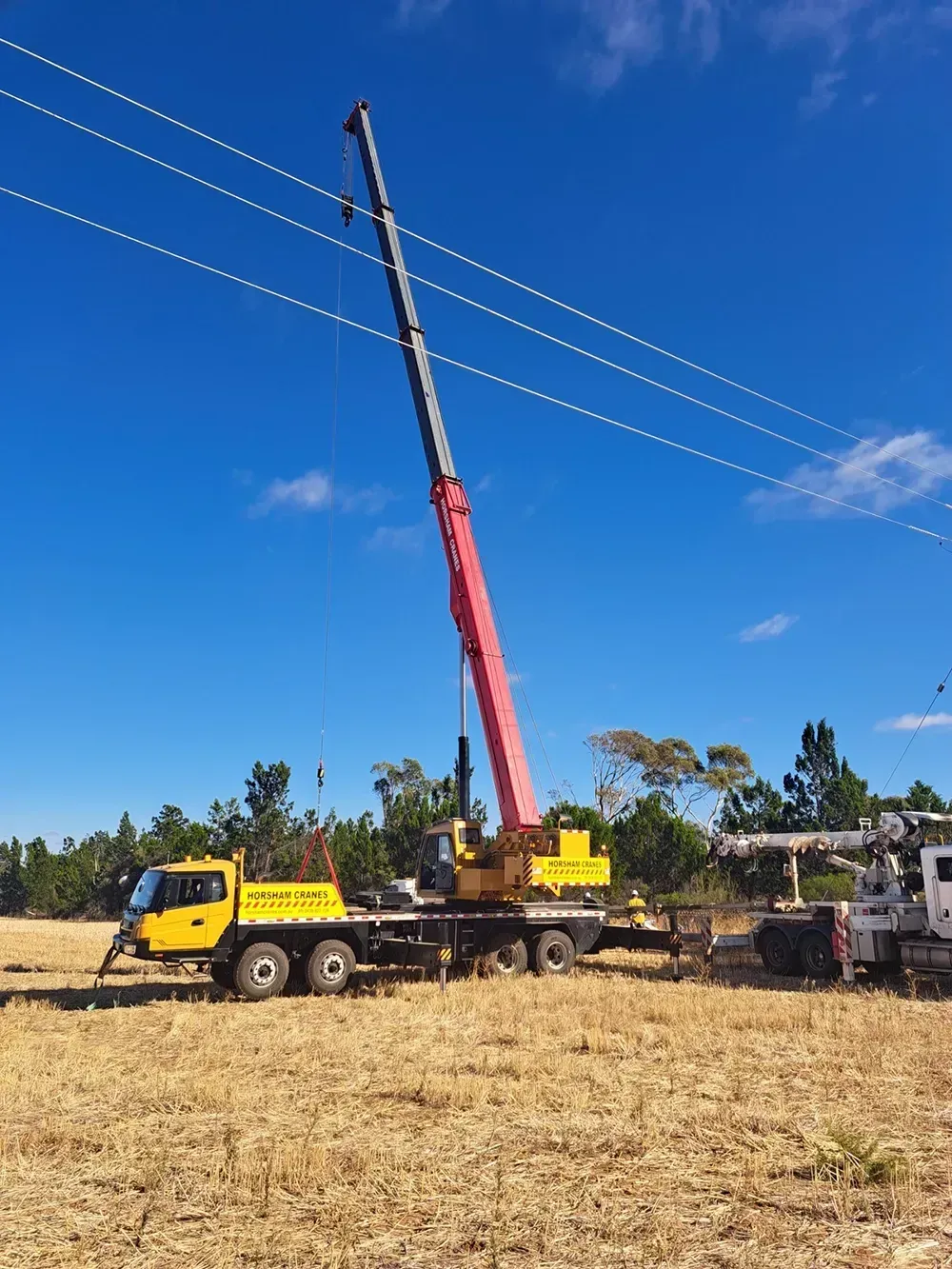 Yellow crane truck with extended red boom near power lines in a field under a blue sky.