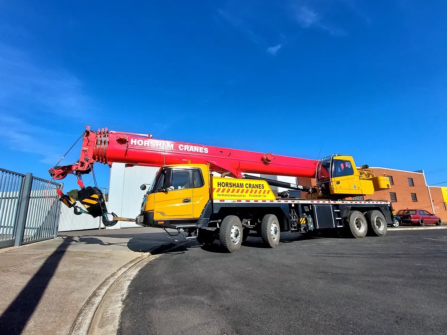 A yellow crane with a red boom is parked on a black surface. The crane is under a blue sky.