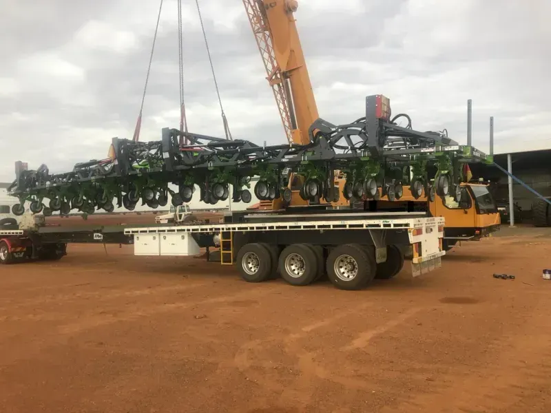 Crane lifting large agricultural planter from a flatbed truck in an outdoor setting.
