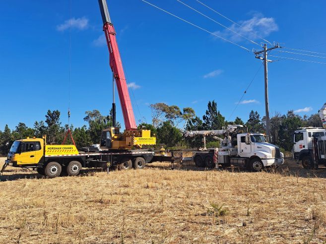 Yellow crane truck lifts something near power lines, white truck nearby, field, blue sky.