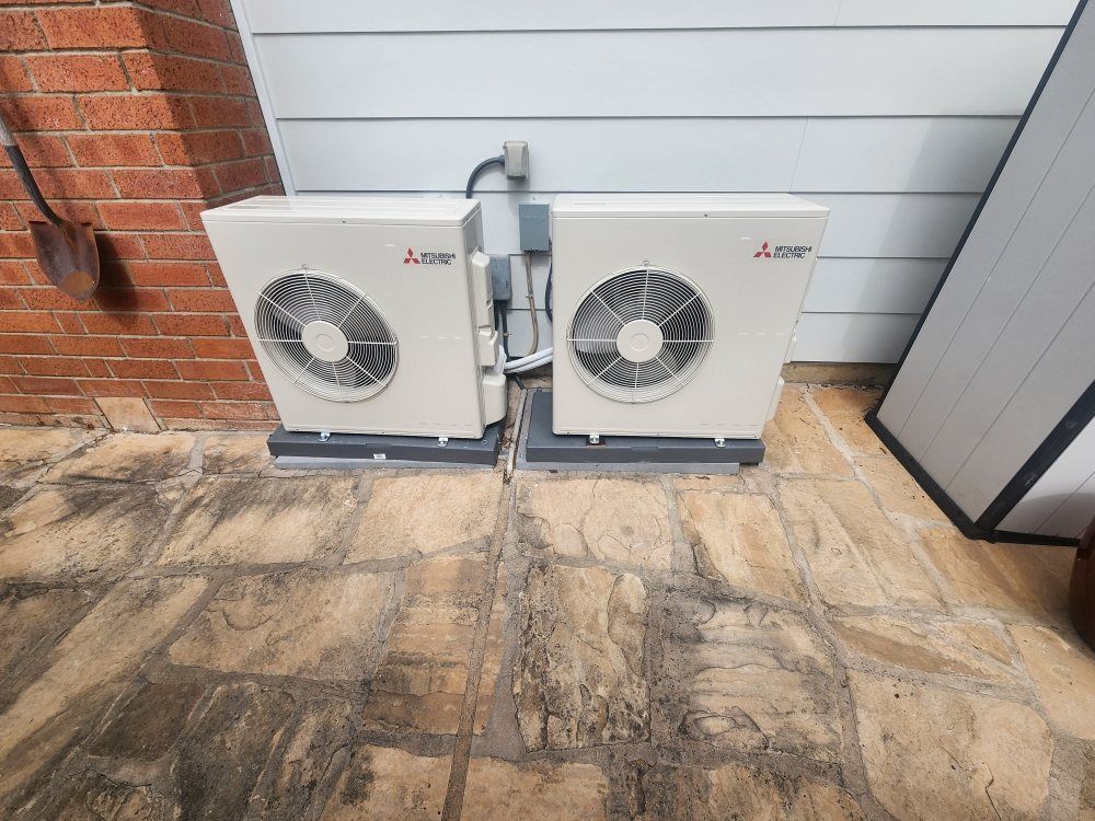 Two white outdoor HVAC units beside a brick wall and house siding on a concrete patio.