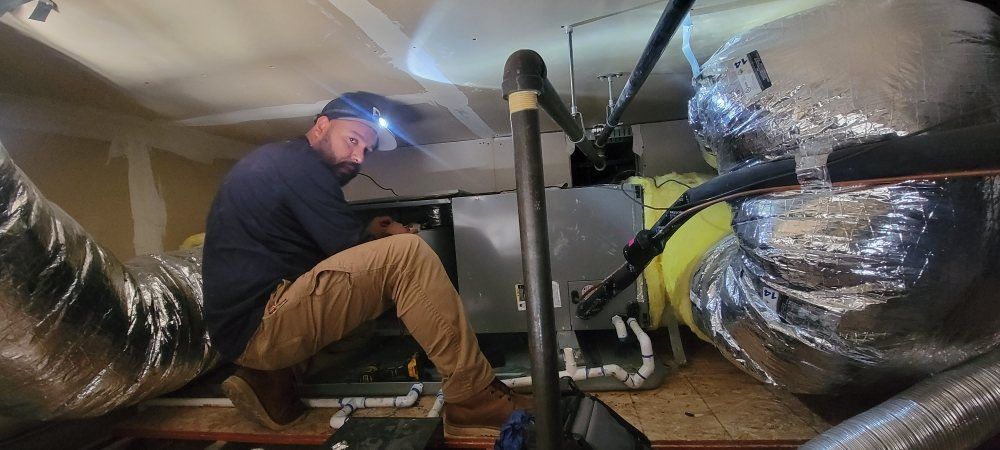 Technician inspecting HVAC ducts in a crawl space with flashlight and insulated silver ductwork