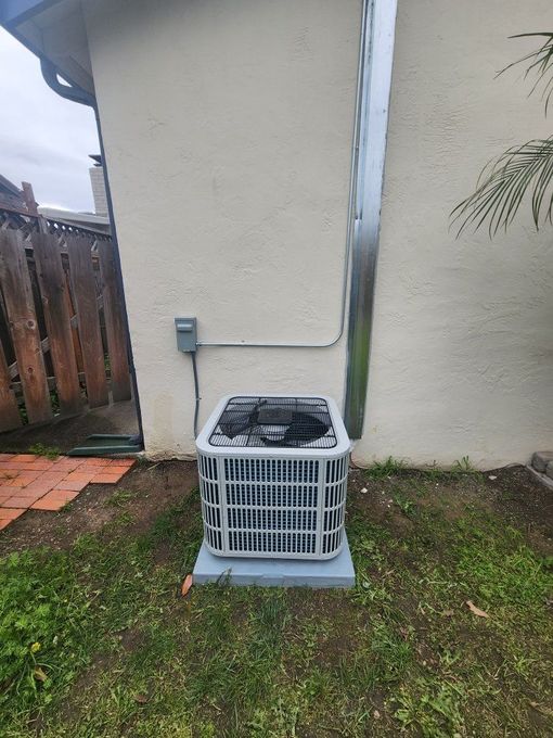 Outdoor air conditioner unit beside a beige house wall, next to a wooden fence and grass.