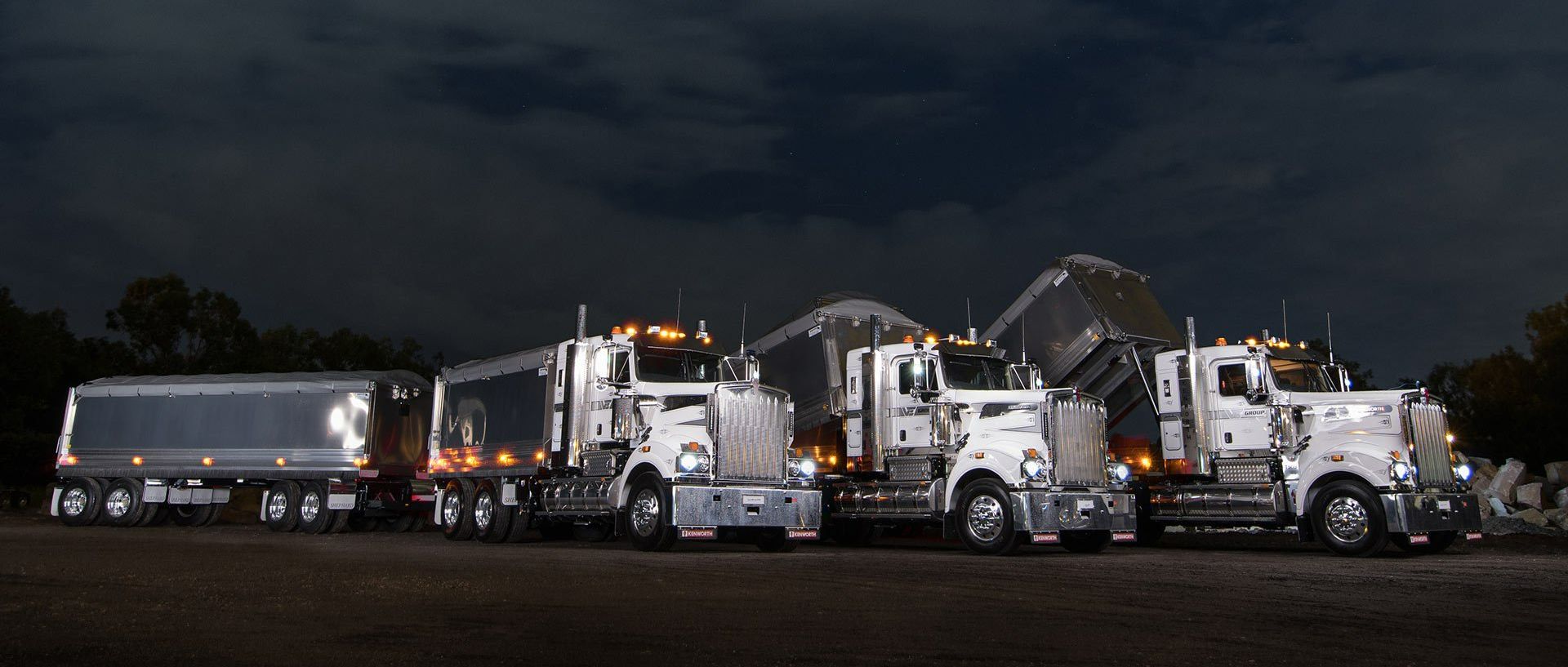 A row of semi trucks are parked in a lot at night.