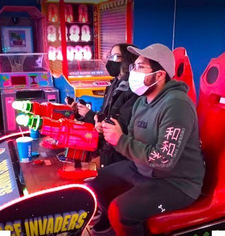 Two people wearing masks are playing a video game in an arcade.