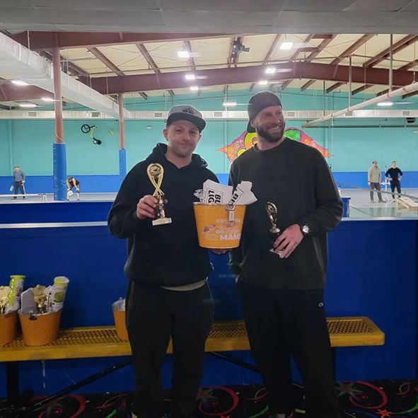 Two men are standing next to each other in a roller rink holding trophies.
