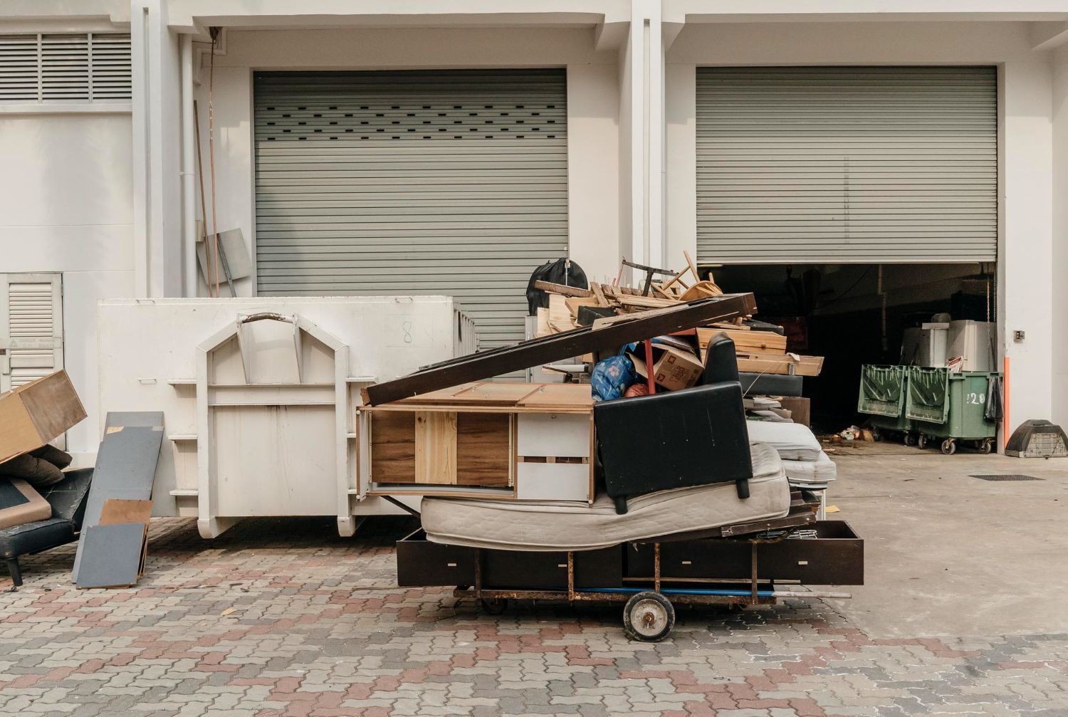 A metal cart overflowing with furniture and debris next to a dumpster outside a building with closed garage doors.