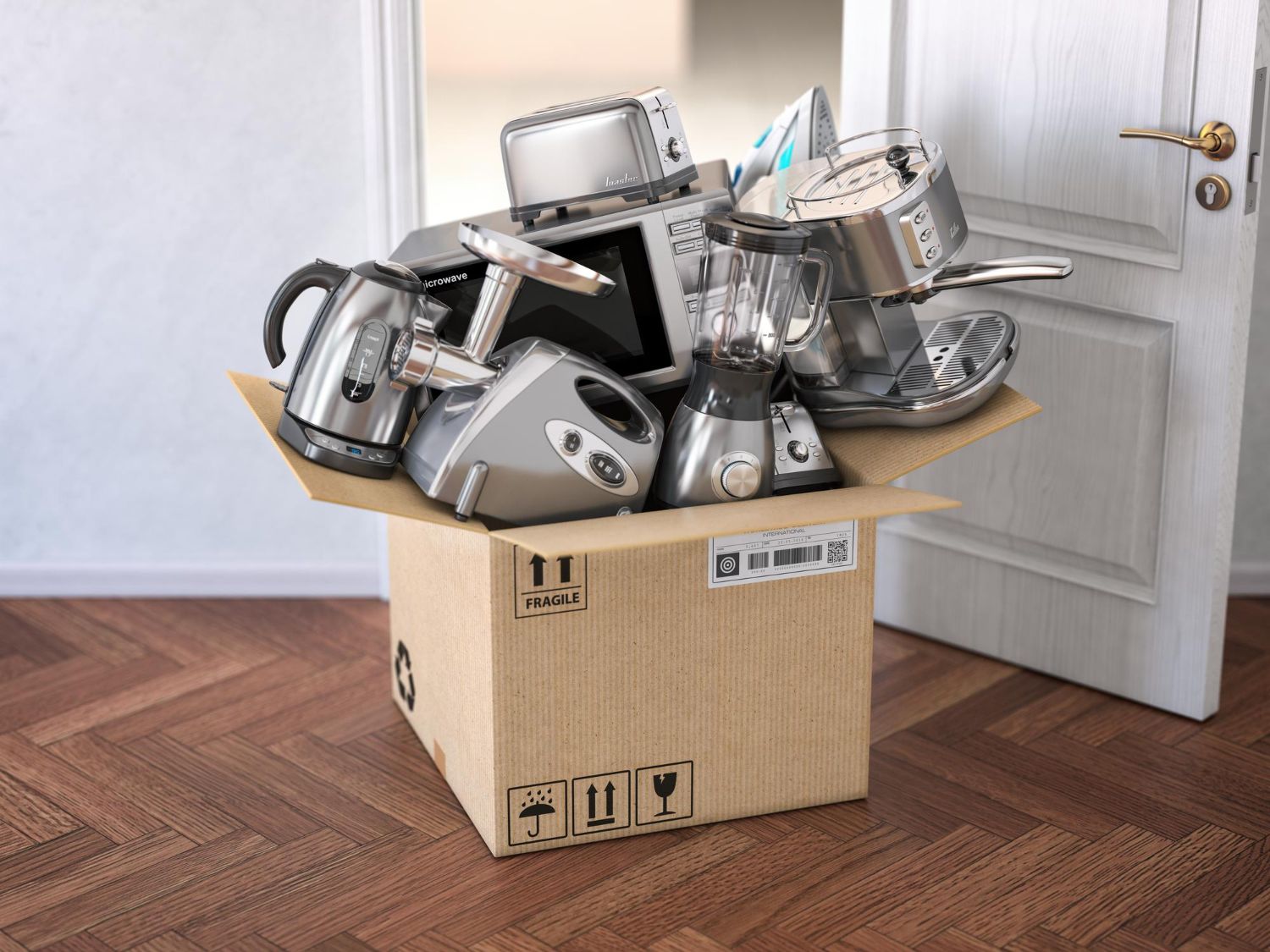 Cardboard box overflowing with various kitchen appliances on a wooden floor near a doorway.