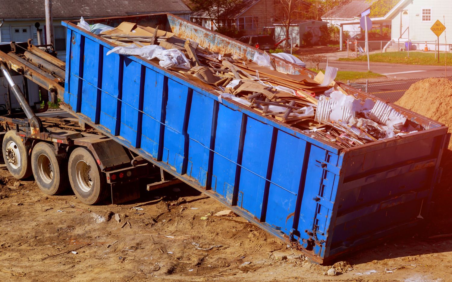 Blue dumpster being tilted to dump debris onto a brown dirt pile.