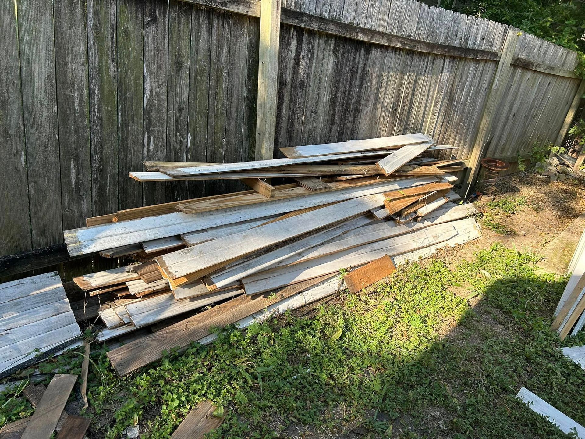 Pile of weathered wooden planks on grass next to a wooden fence.