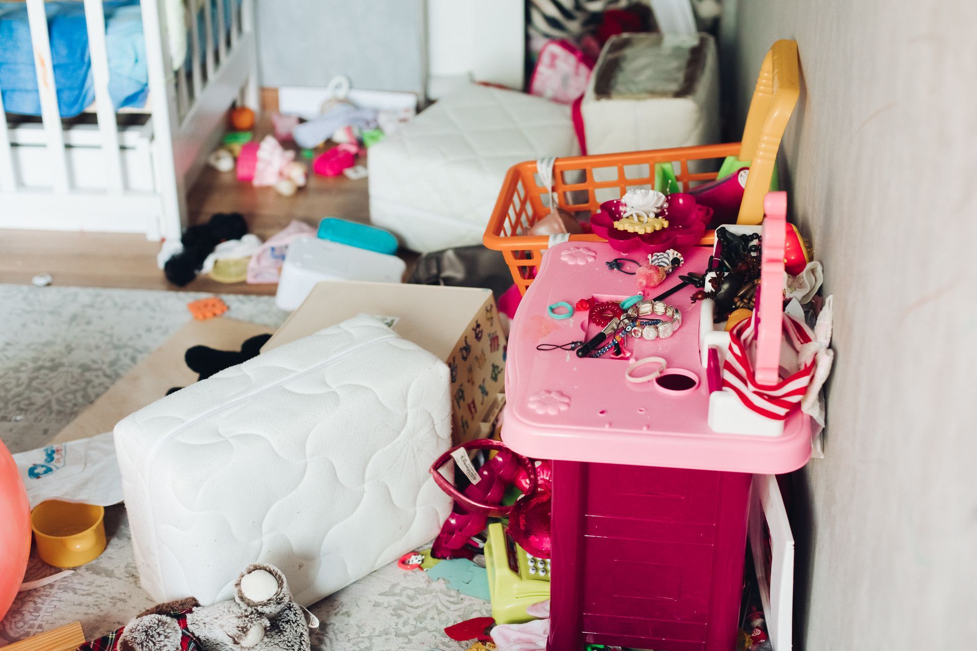 A messy room with toys scattered around a pink toy desk, a crib, and a white cushioned box.