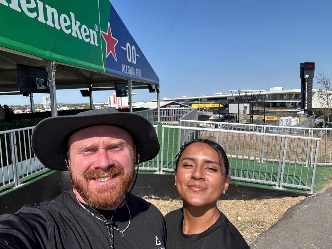 Two people smiling for a selfie in front of a Heineken tent and stadium.