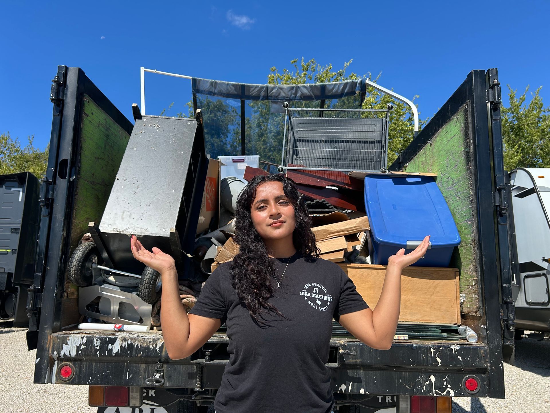 Woman stands with hands up in front of a truck bed filled with junk on a sunny day.