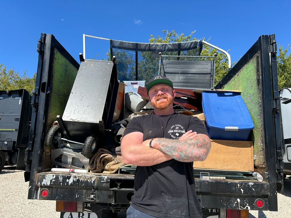 Man with arms crossed, stands in front of a truck bed filled with scrap metal and other items.