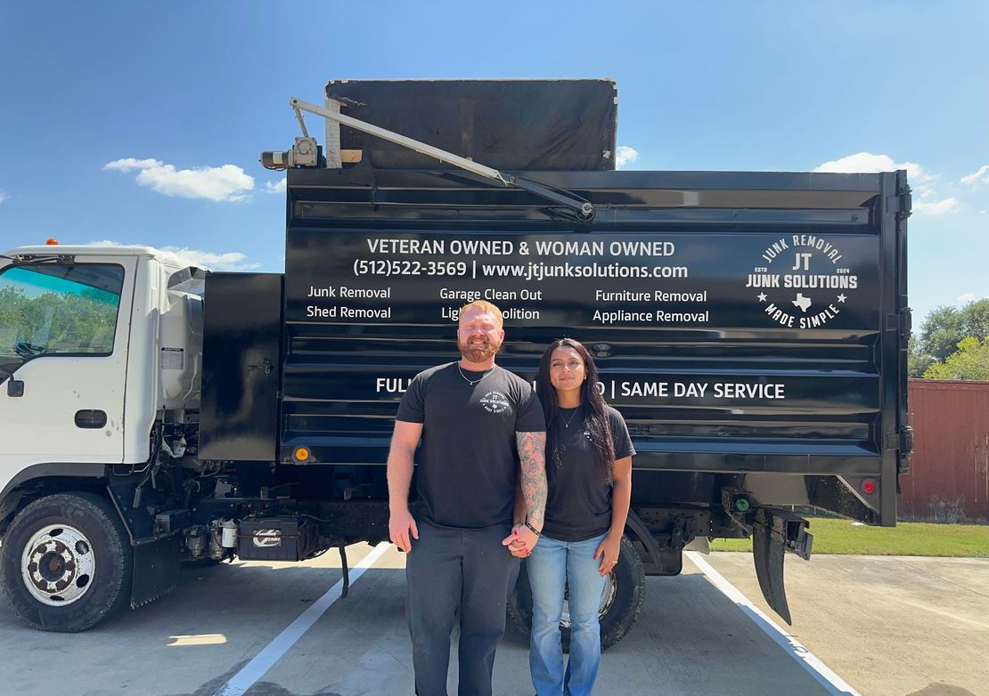 A man and woman stand in front of a black dump truck with business lettering, outside on a sunny day.