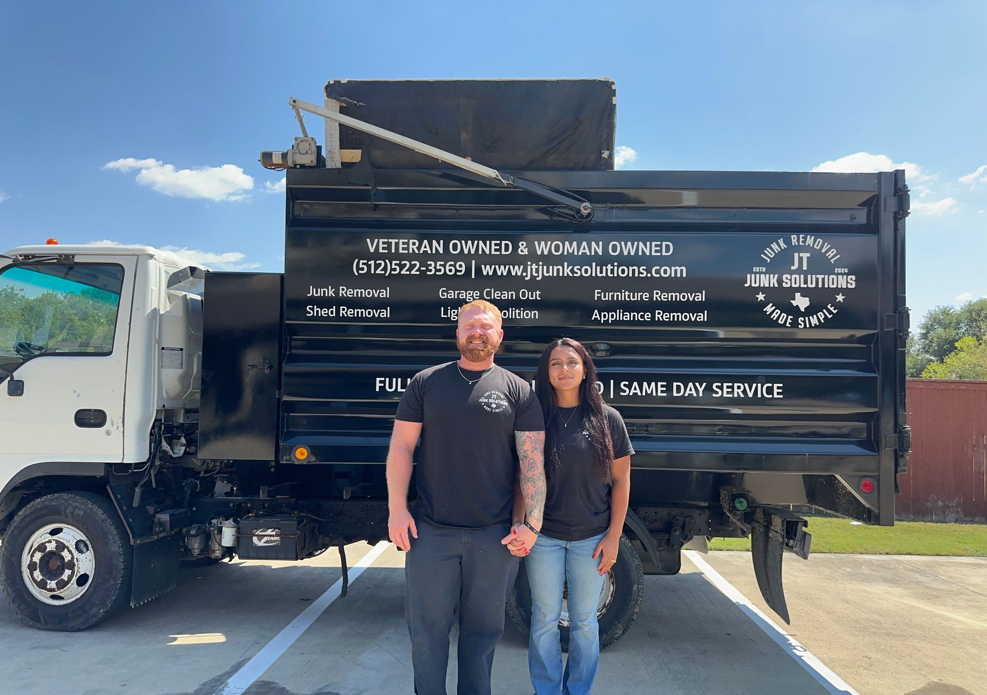 A man and woman stand in front of a black dump truck with business lettering, outside on a sunny day.