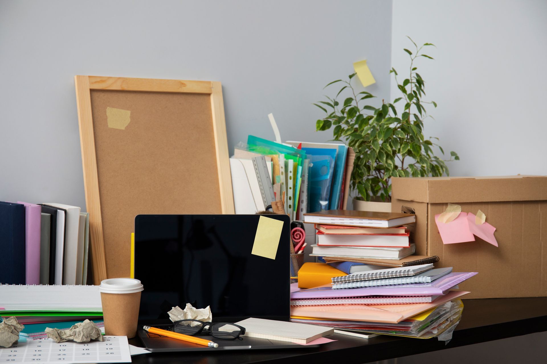 A messy desk with a laptop , books , papers , and a cardboard box.