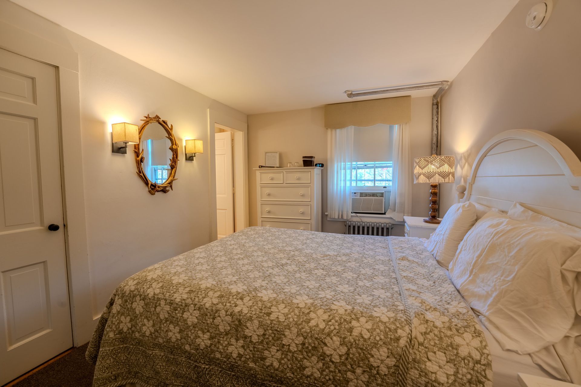 Bedroom with white bed, patterned quilt, gold mirror, and dresser. Window with air conditioner.